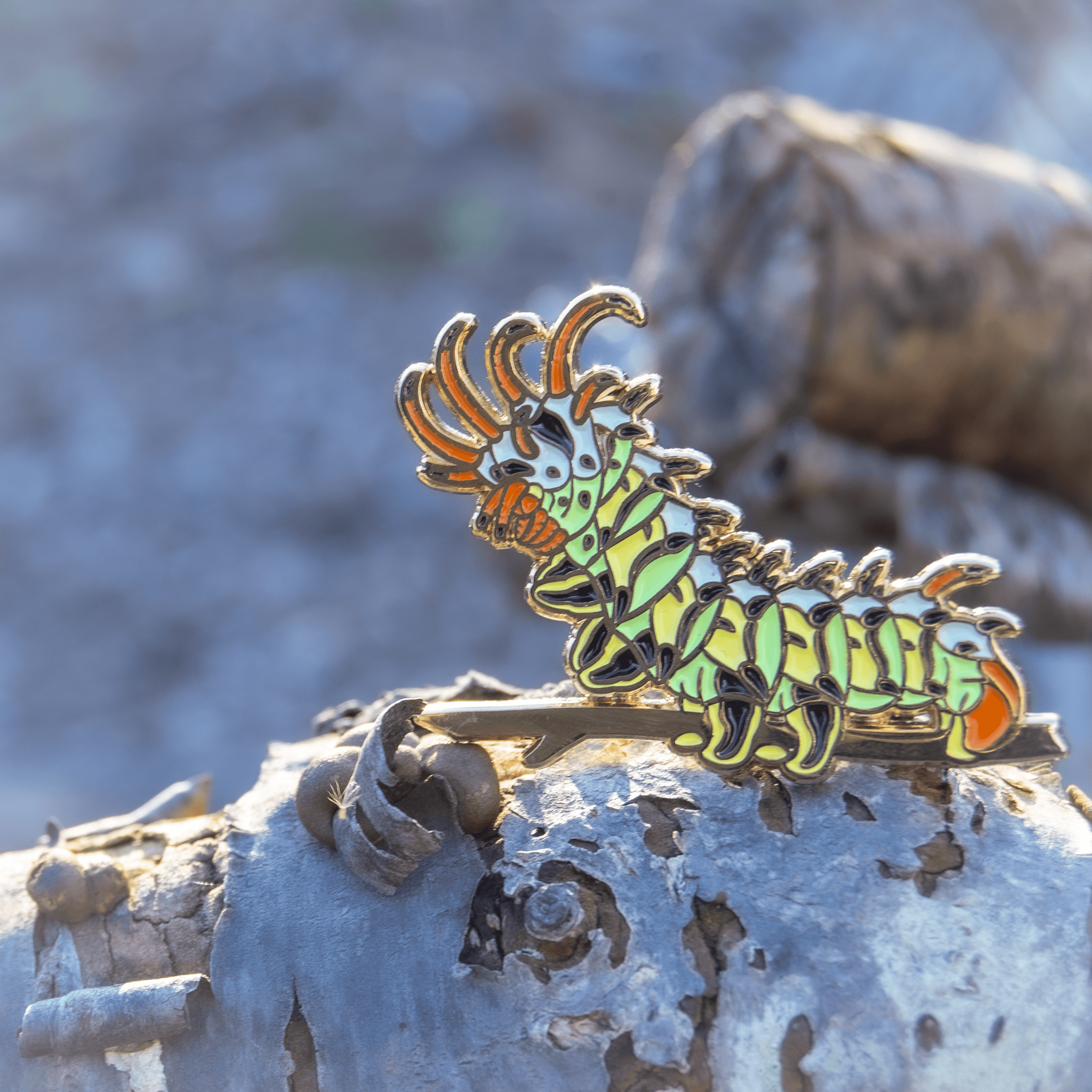 A green, yellow, black, orange, blue, and gold enamel pin of the Citheronia Regalis caterpillar, the Hickory Honrned Devil, placed upon a log and sparkling in the daylight.