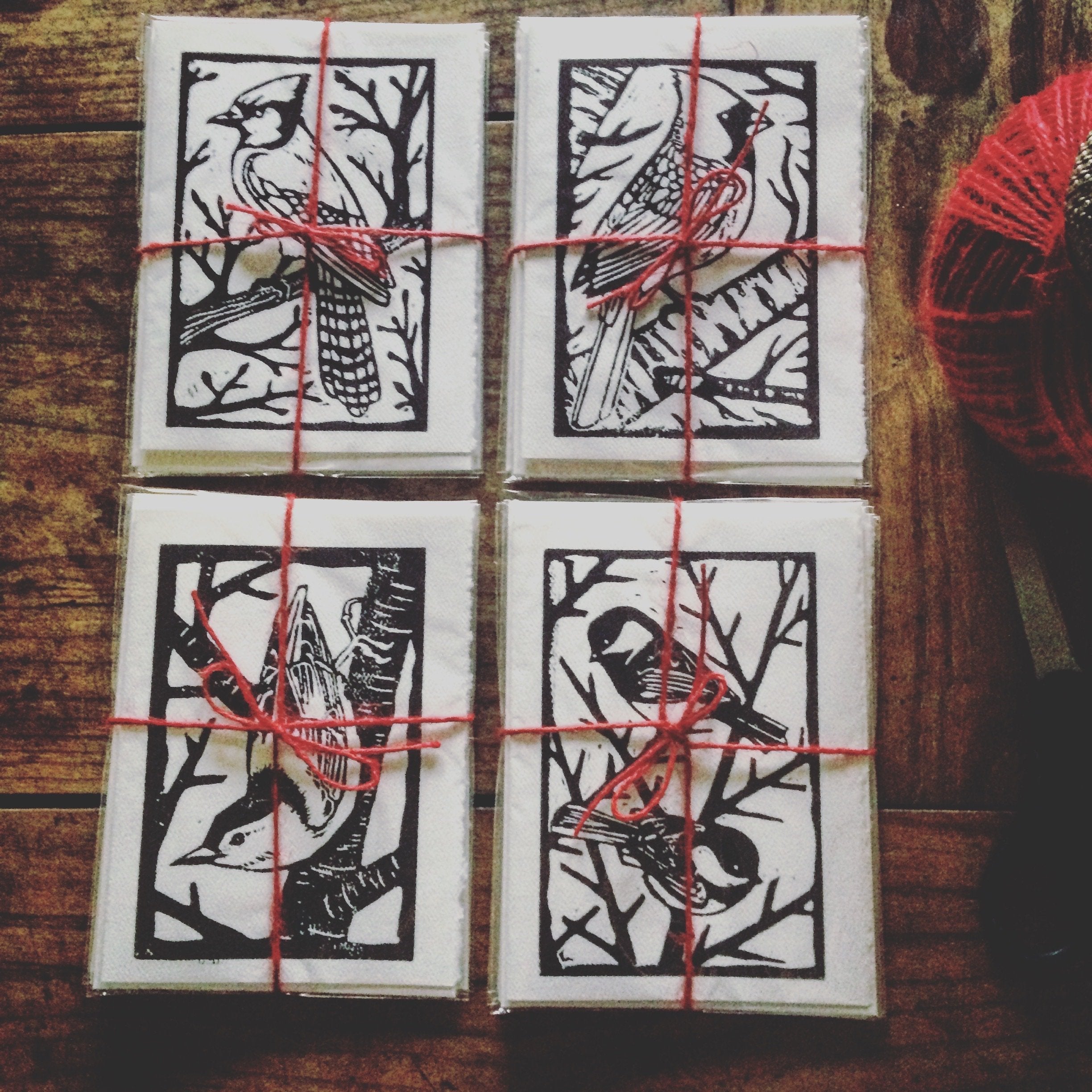 Hand-made block printed cards of birds laid out on a wooden table, including a bluejay, cardinal, nuthatch, and chickadee.