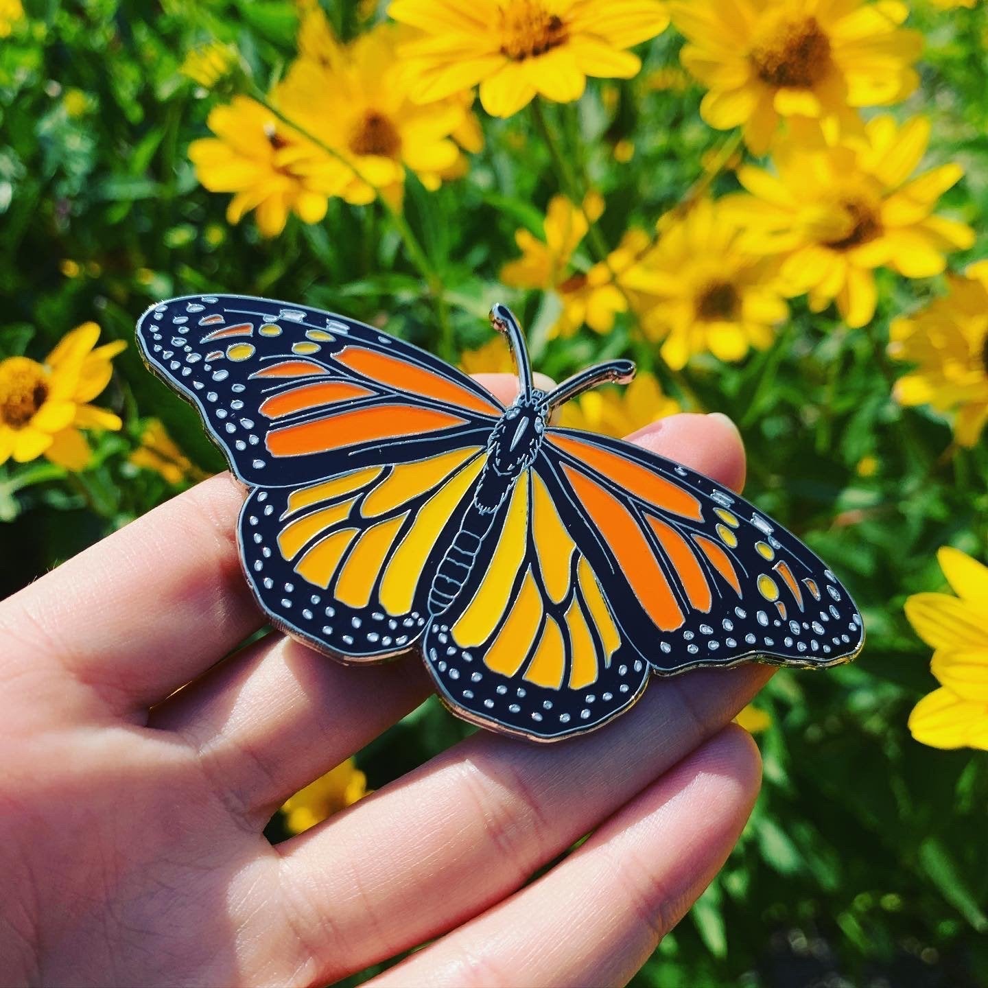 A black, orange, and white monarch butterfly (Danaus plexippus) enamel pin being held in a person's hand above yellow flowers.