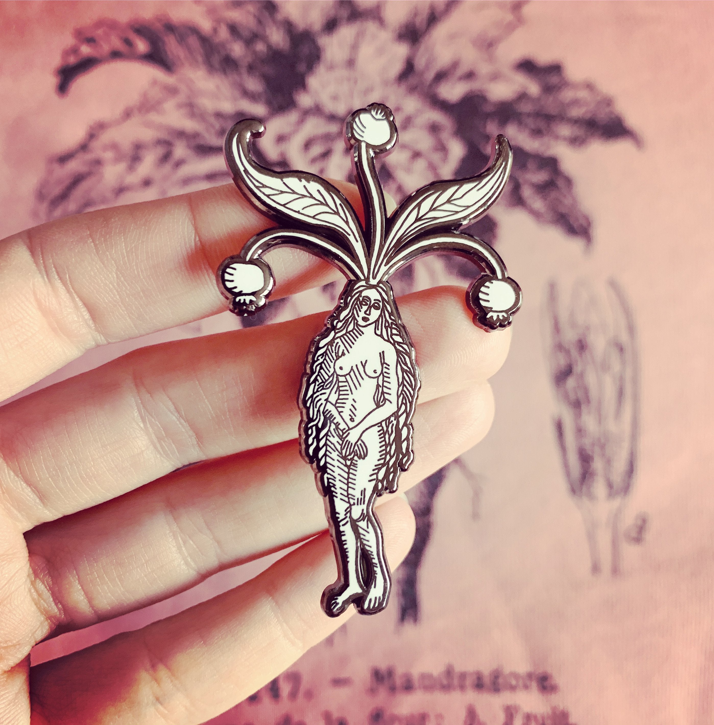 A black and white hard enamel pin of a nude woman, with mandrake leaves and berries growing from her head. The artwork is from a medieval woodcut featured in the Hortus Sanitatis encyclopedia. In the background is a pink shirt with a mandrake botanical plate.